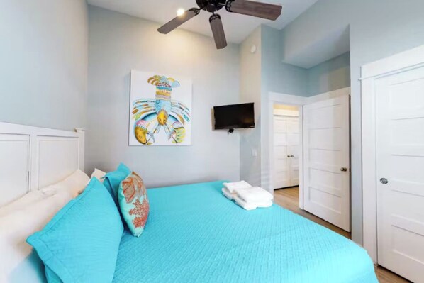 A bedroom with a bed, a ceiling fan, a TV, and a door leading to another room.