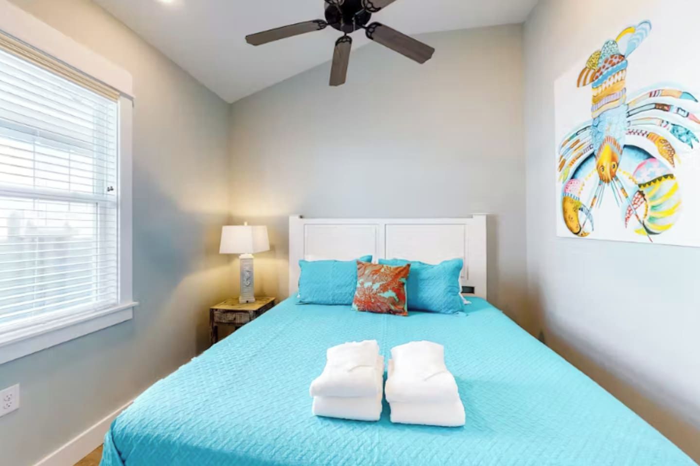 A bedroom with a bed, a ceiling fan, a window with blinds, a nightstand with a lamp, and a wall art piece.