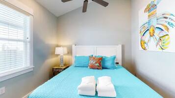 A bedroom with a bed, a ceiling fan, a window with blinds, a nightstand with a lamp, and a wall art piece.