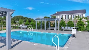 A swimming pool with a pergola and lounge chairs, surrounded by greenery and a building in the background.