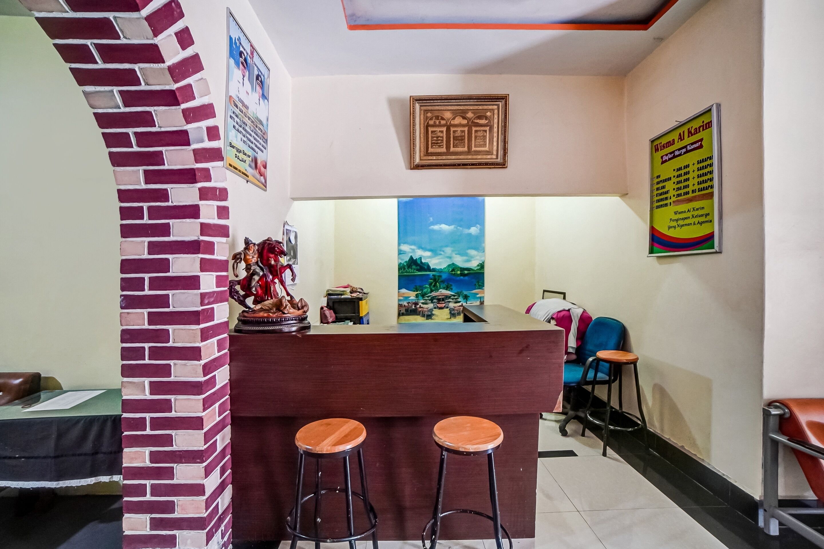 A reception area with a brick pillar, wooden counter, two stools, a painting, and a poster on the wall.