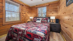 A wooden cabin bedroom with a bed, two bedside tables, a ceiling fan, and windows with blinds.