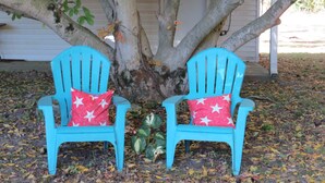 Zwei türkisfarbene Adirondack-Stühle mit roten Kissen mit weißen Sternen, platziert unter einem Baum mit Herbstlaub auf dem Boden.
