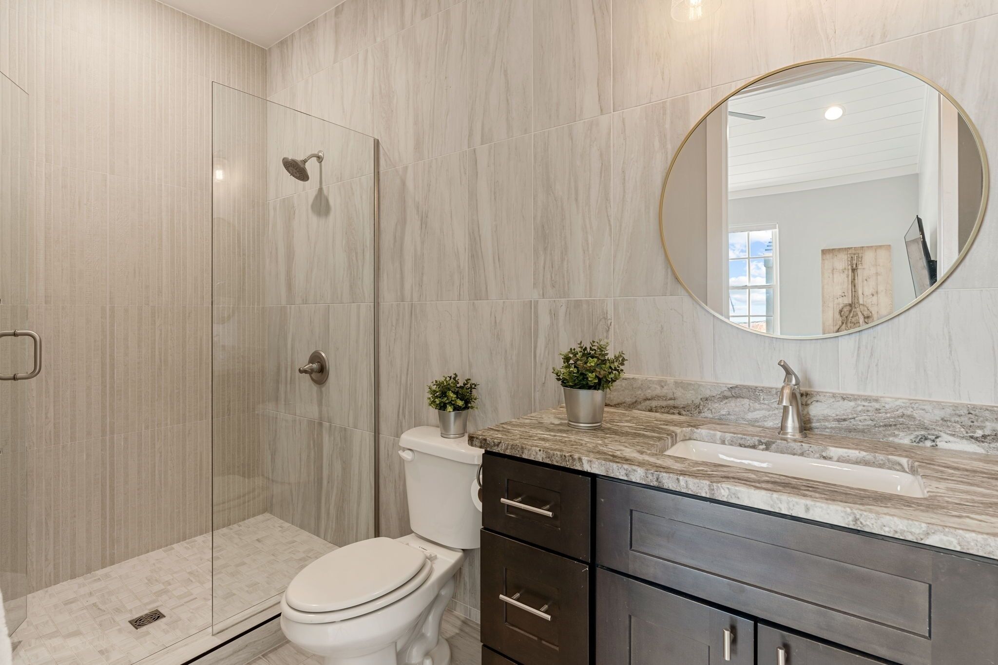 Un baño moderno con cabina de ducha de vidrio, inodoro, lavamanos con encimera de mármol y mueble de tocador de madera oscura.