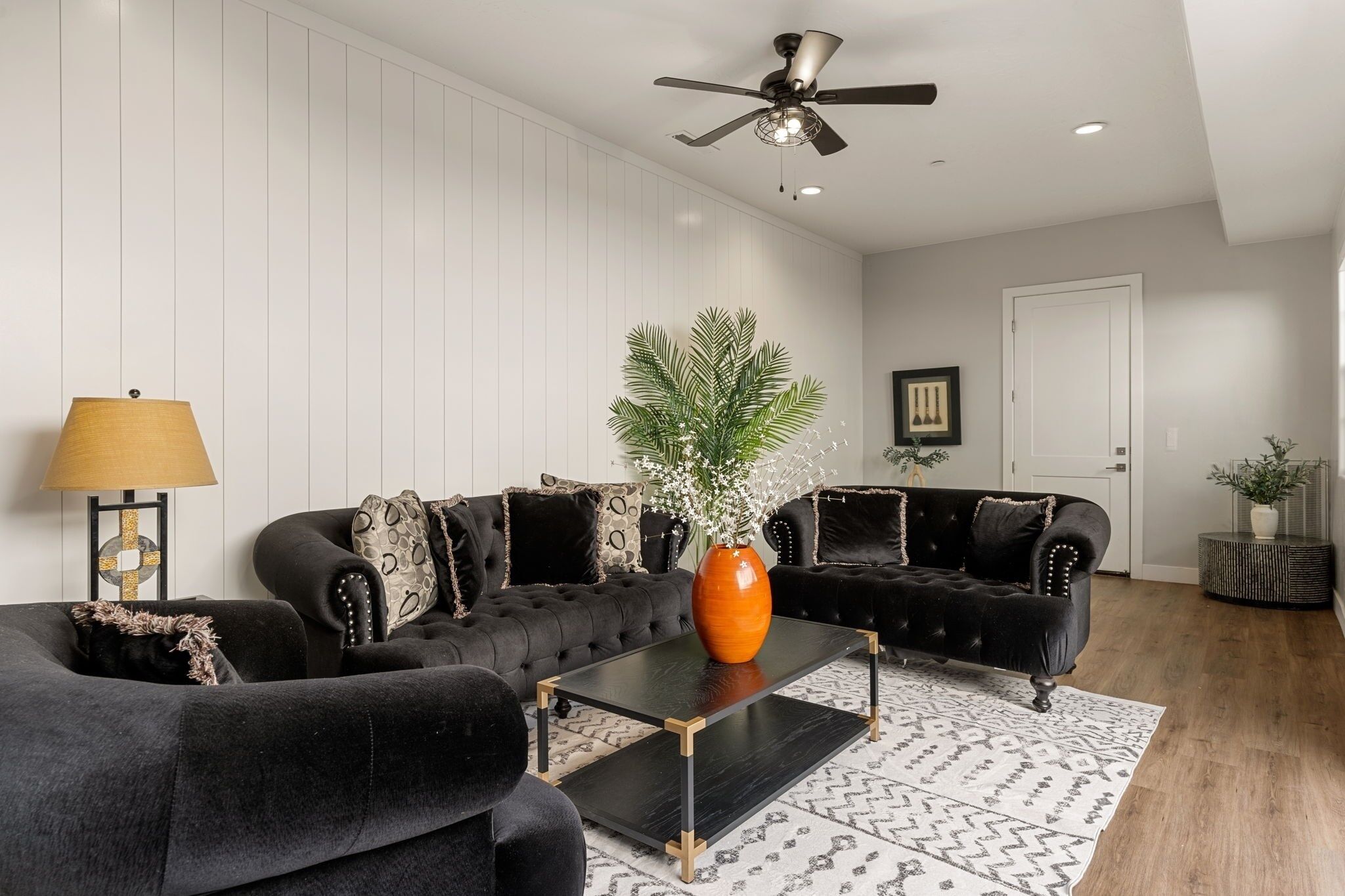 A modern living room with a black sofa set, a glass coffee table, and a ceiling fan.