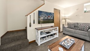 Uma sala de estar com escada, um sofá, uma mesa de centro com conchas e uma televisão na parede, além de um quadro com a palavra "LOVE".
