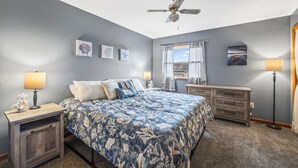 Quarto com cama, mesas de cabeceira, cómoda, ventoinha no teto e obras de arte nas paredes.