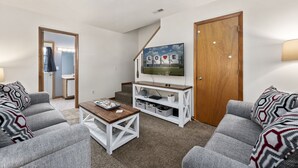 Sala de estar com sofás cinzas, uma mesa de centro, uma televisão sobre um suporte branco e uma porta de madeira.