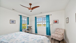 Une chambre à coucher comprenant un lit, une chaise, un bureau, deux fenêtres avec des rideaux, un ventilateur de plafond et des tableaux encadrés accrochés aux murs.