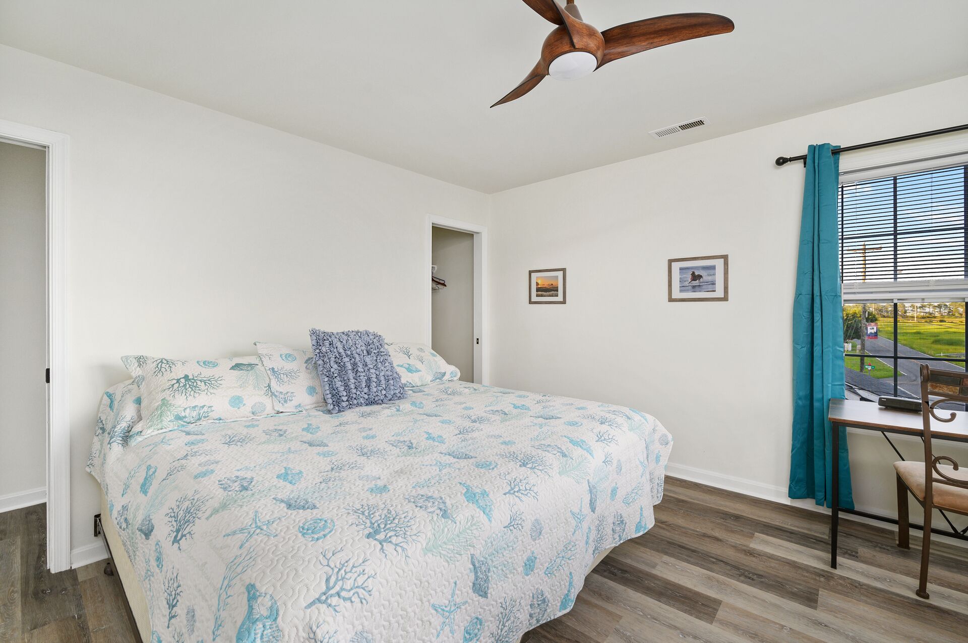 Un dormitorio con cama, ventilador de techo, ventana con cortinas y una pequeña mesa con una silla.
