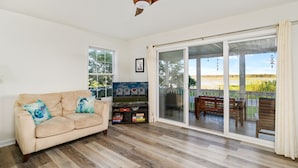 Un salon avec un canapé beige, un plancher en bois, un ventilateur de plafond, un téléviseur et une porte-fenêtre coulissante donnant sur une terrasse extérieure avec une plateforme en bois et une vue sur un champ.