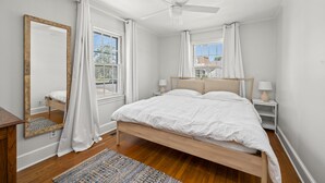 A bedroom with a wooden bed, white bedding, a ceiling fan, two bedside tables with lamps, a large mirror, and a view of the outside through a window.