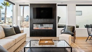A modern living room with a flat-screen TV, a fireplace, a sofa, and a dining area with chairs.
