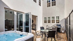 Ein Whirlpool mit Glasumrandung, ein Esstisch mit Stühlen und ein Grill vor einem Gebäude mit großen Fenstern.