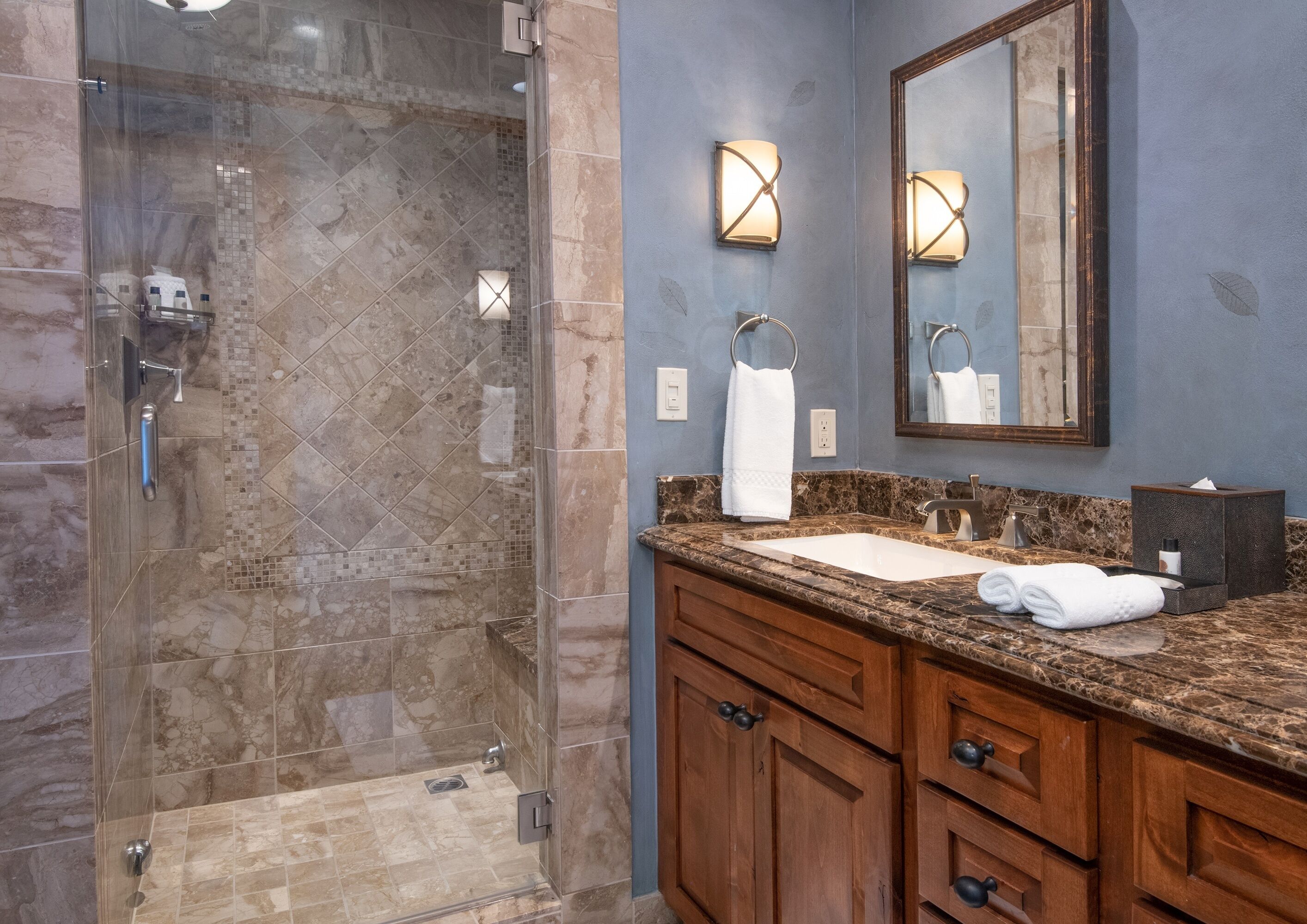 A modern bathroom with a glass-enclosed shower, marble tiles, a sink with a marble countertop, wooden cabinets, and a large mirror.