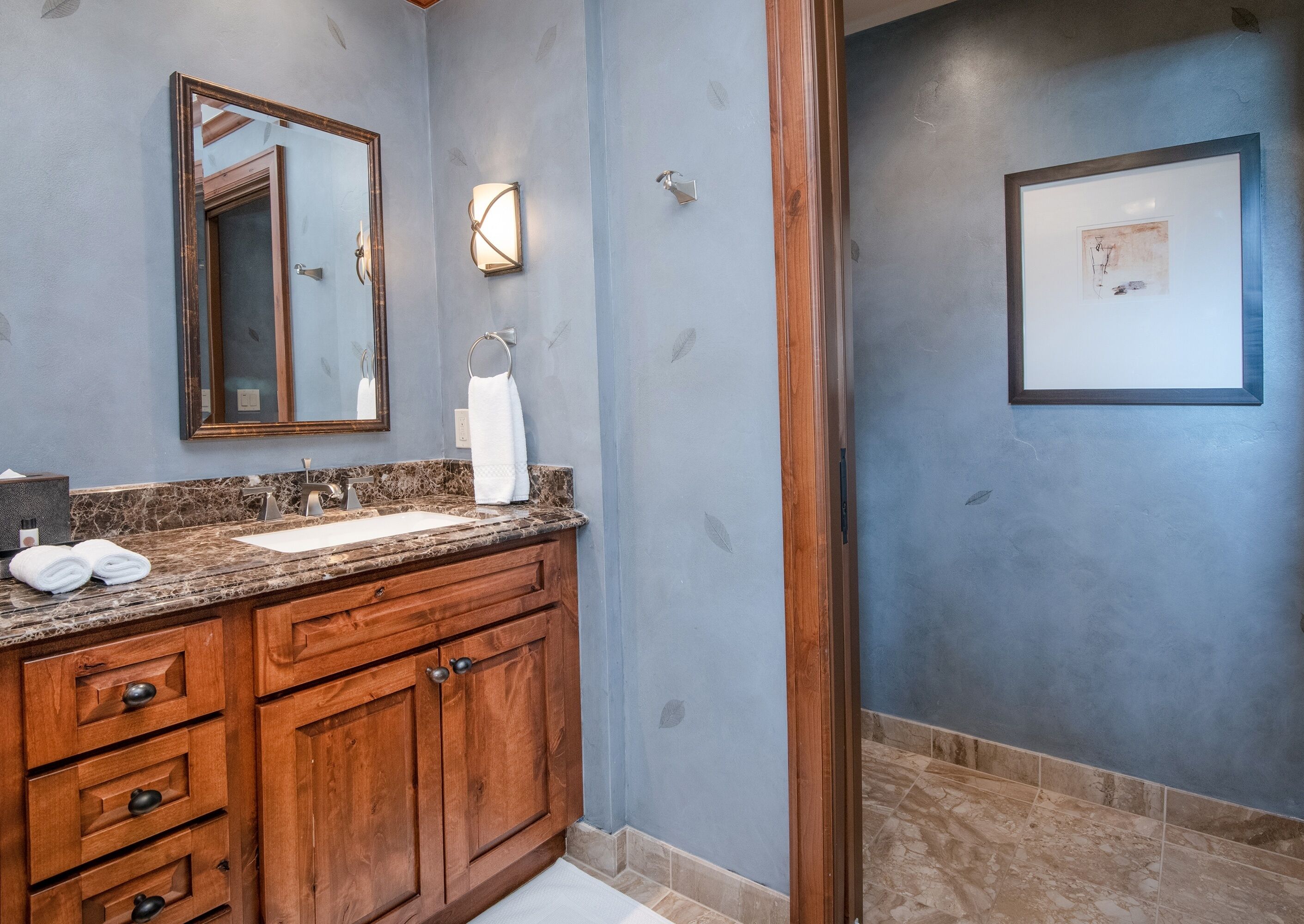 Un baño con encimera de mármol, mueble de tocador de madera, un espejo, toallero y una obra de arte enmarcada en la pared.