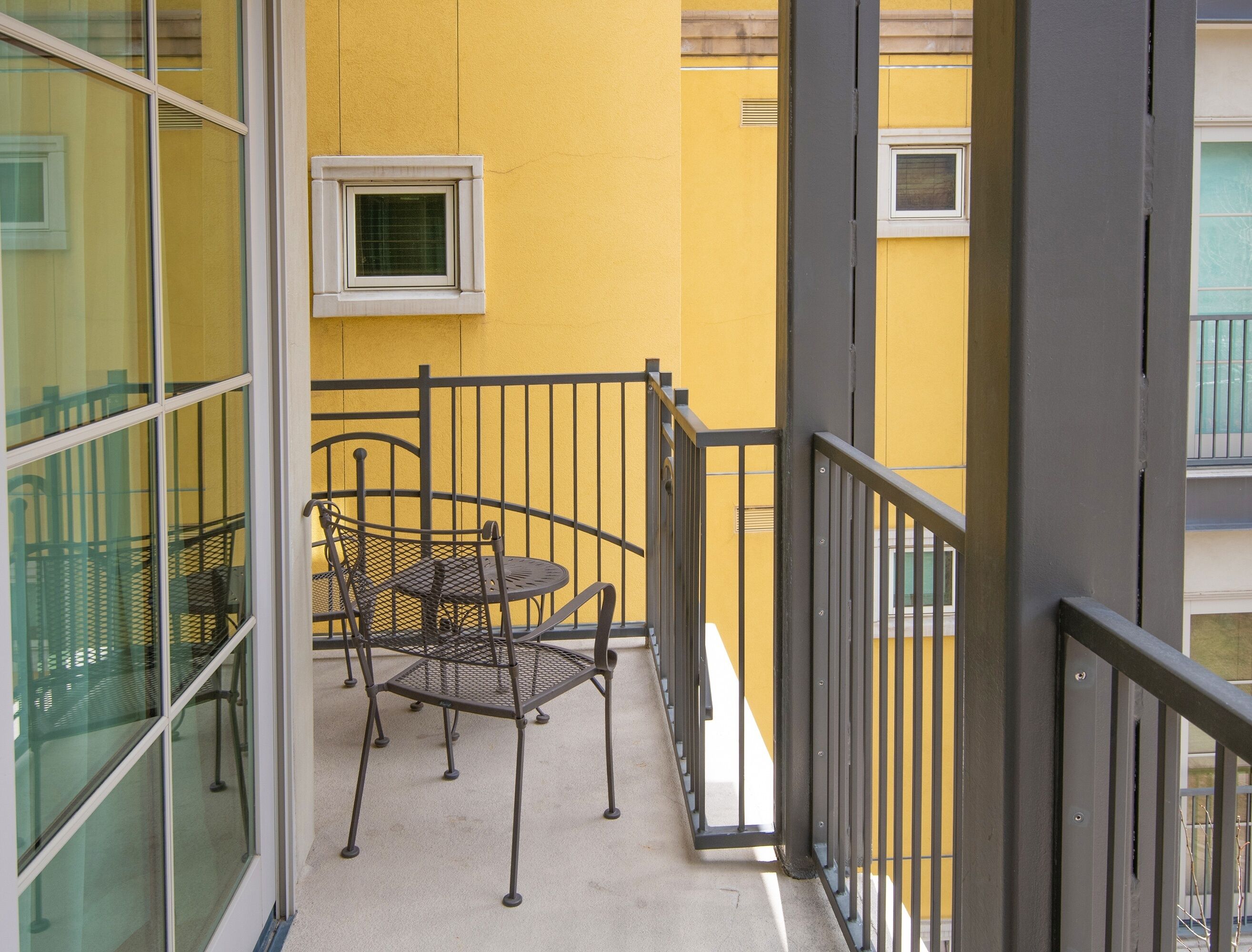 Ein Balkon mit einem Metalltisch und Stühlen, einem kleinen Fenster und einer gelben Wand.