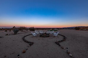 Un paysage désertique avec une disposition circulaire de chaises blanches autour d’un foyer central, le tout se détachant sur le fond d’un coucher de soleil sur des collines lointaines.