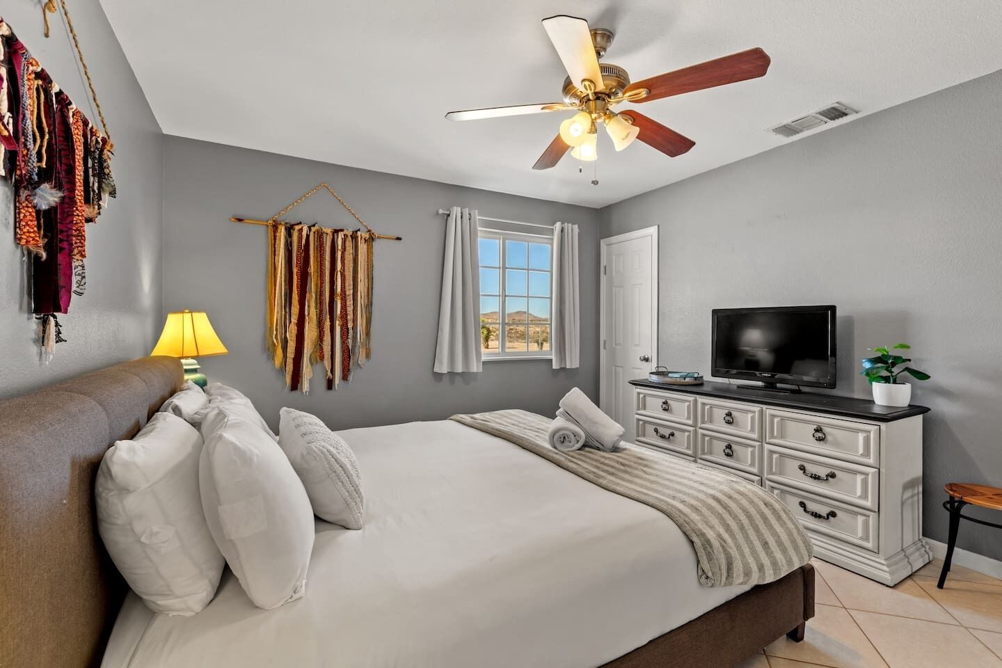 A bedroom with a bed, a TV, a dresser, a ceiling fan, and a window with curtains.