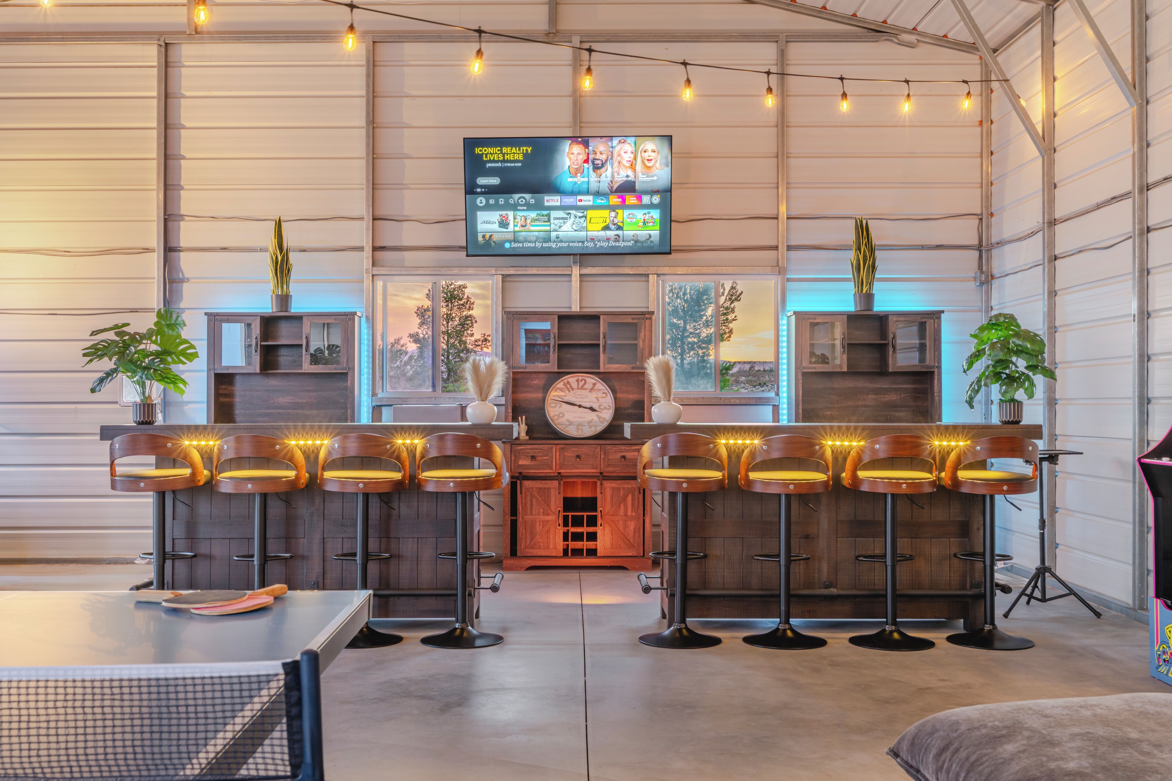 Zona de bar con una pantalla de televisión, taburetes de madera y plantas en macetas.