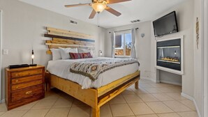 A bedroom with a wooden bed, a ceiling fan, a TV mounted on the wall, and a fireplace.