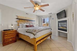 A bedroom with a wooden bed, a ceiling fan, a TV mounted on the wall, and a fireplace.