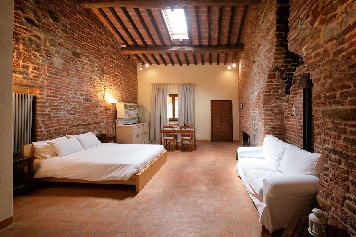 Ein Schlafzimmer mit Backsteinwand, Holzbalkendecke, einem Bett, einem weißen Sofa, einem Tisch und einem Stuhl.