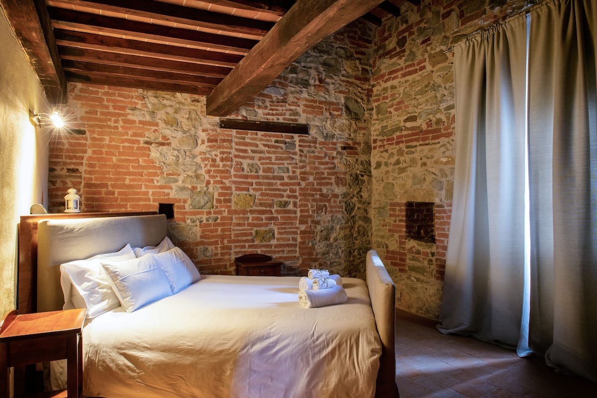 Ein Zimmer mit Backsteinwand, Holzboden und einem Bett mit weißer Bettwäsche sowie einem Nachttisch mit Lampe.
