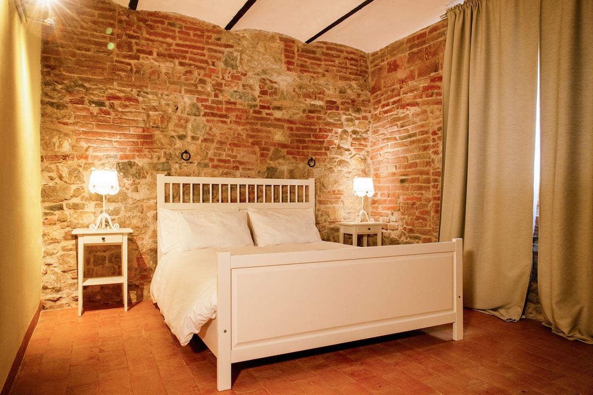 Ein Zimmer mit Backsteinwand, einem weißen Bett, zwei Nachttischen mit Lampen und einem Vorhang.
