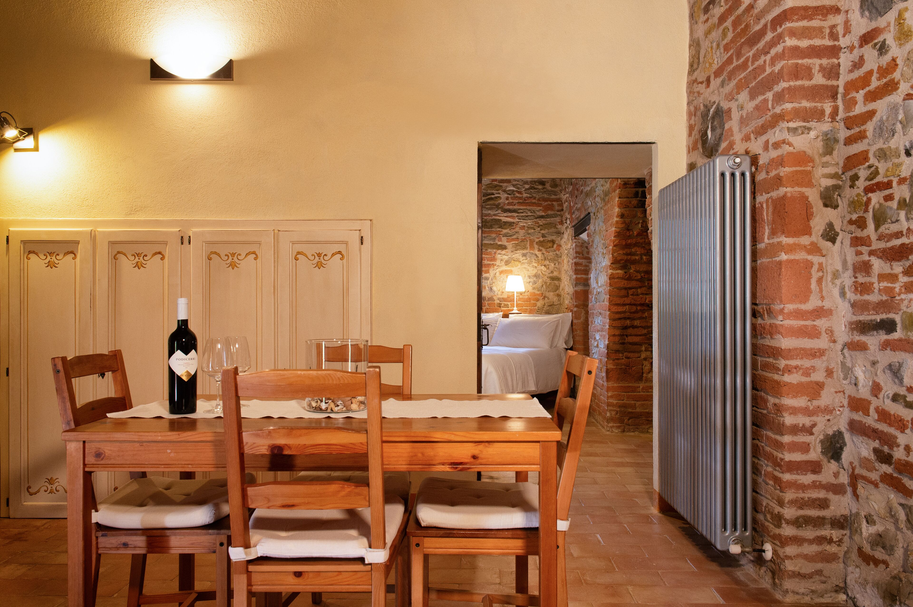 Un comedor con una mesa de madera, sillas y una botella de vino. Se aprecia una pared de ladrillo y un radiador al fondo.