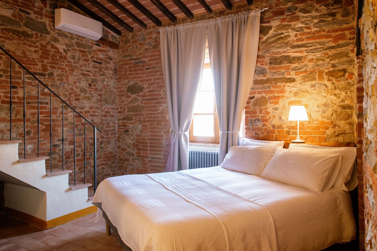 Ein Schlafzimmer mit einer Steinsäule, einem Bett mit weißer Bettwäsche, einem Nachttisch mit einer Lampe und einer Treppe mit Metallgeländer.