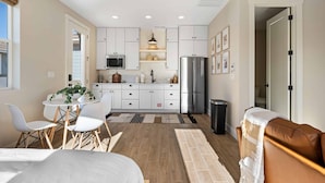Cocina compacta con armarios blancos, un refrigerador de acero inoxidable y un microondas. Hay una zona de comedor con una mesa y sillas, y un sofá de cuero marrón.