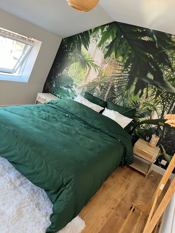A bedroom with a large bed, a nightstand, a wooden floor, and a wall mural of tropical foliage.