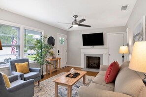 A living room with a fireplace, a ceiling fan, a television, and a window with blinds. - Central Midtown Cottage (Mobile)