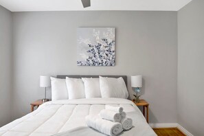 A neatly made bed with white bedding and rolled towels, a gray headboard, and a wall-mounted artwork.