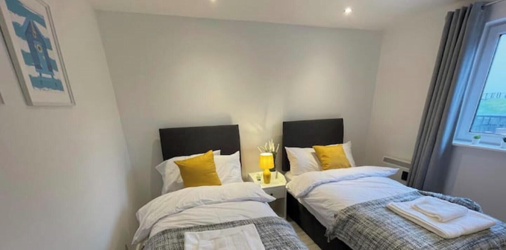 Ein Doppelzimmer mit zwei Betten, jeweils mit Kopfteil, weißer Bettwäsche und einem aufgebetteten Handtuch am Fußende. Zwischen den Betten befindet sich ein Nachttisch mit einer Lampe.