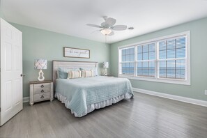 A bedroom with a bed, a nightstand, a ceiling fan, a window with blinds, and a door. - Southern Belle: Oceanfront | Surfside Beach Vacation Rental (Surfside Beach)