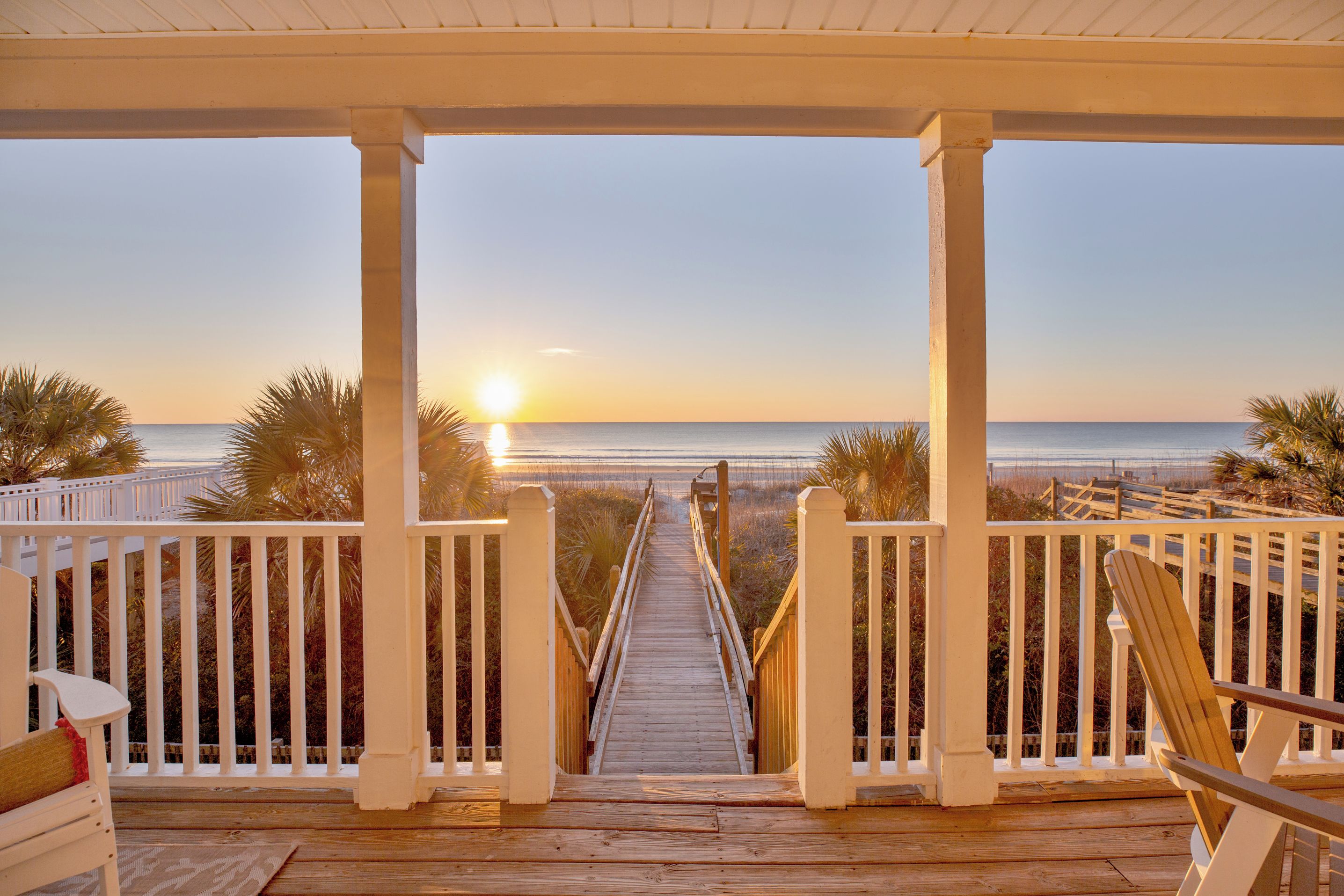 A wooden deck with a view of the sunset over the ocean, white railings, and a chair.