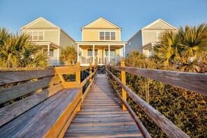 A wooden boardwalk leading to a row of two-story beach houses with a covered porch and a clear blue sky.