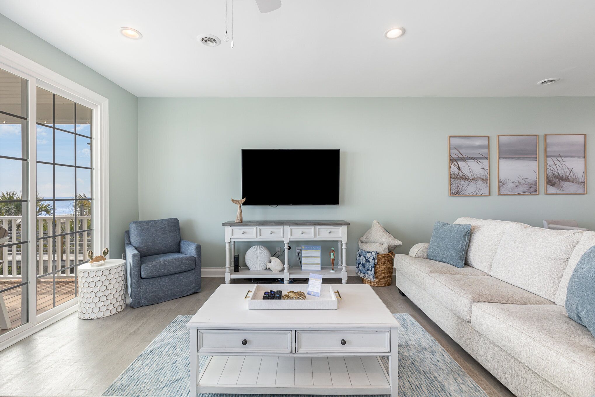 A living room with a white coffee table, a grey armchair, a white sofa, and a flat-screen TV mounted on the wall.