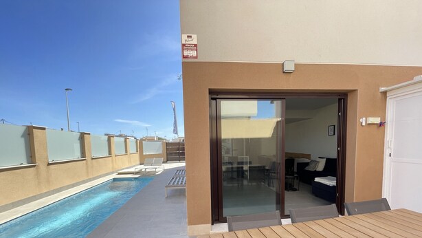 Ein Poolbereich mit Holzterrasse, einer Glastür zu einem Zimmer und blauem Himmel.