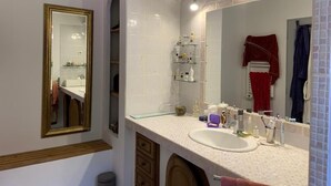 Une salle de bain avec un grand miroir, des murs recouverts de carreaux blancs, un lavabo blanc et des étagères en verre au-dessus du lavabo.