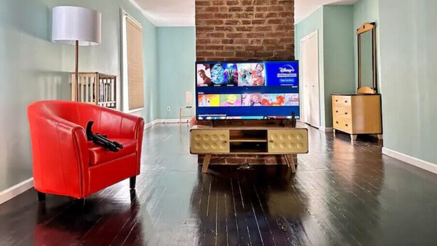 Een woonkamer met een open haard van baksteen, een rode fauteuil, een tv op een houten dressoir en een kaptafel met spiegel.