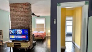 Een kamer met een haard van baksteen, een bed, een televisie en een gang die naar een andere kamer leidt.