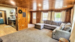 A cozy cabin interior with a wooden ceiling, a gray sectional sofa, a kitchen area, and a door leading to a bathroom. - Lynnhurst Resort-Private cabin on Kentucky Lake with Hot Tub (Murray)