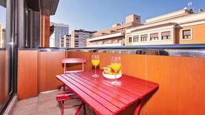 Une table en métal rouge, avec deux verres de jus d’orange et un saladier de fruits, sur un balcon donnant sur la ville.