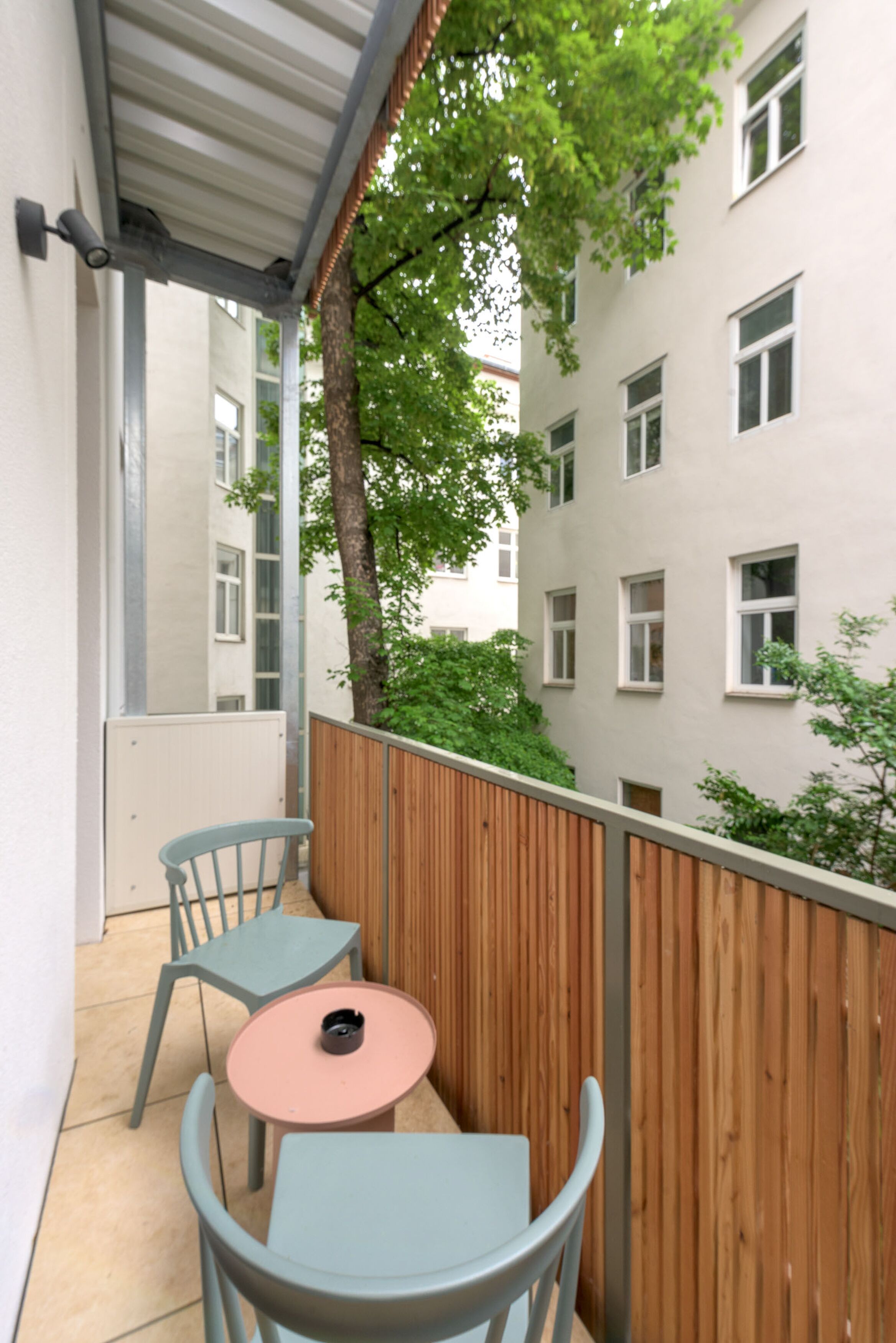 A balcony with two chairs and a small table, a wooden fence, and a building with multiple windows in the background.