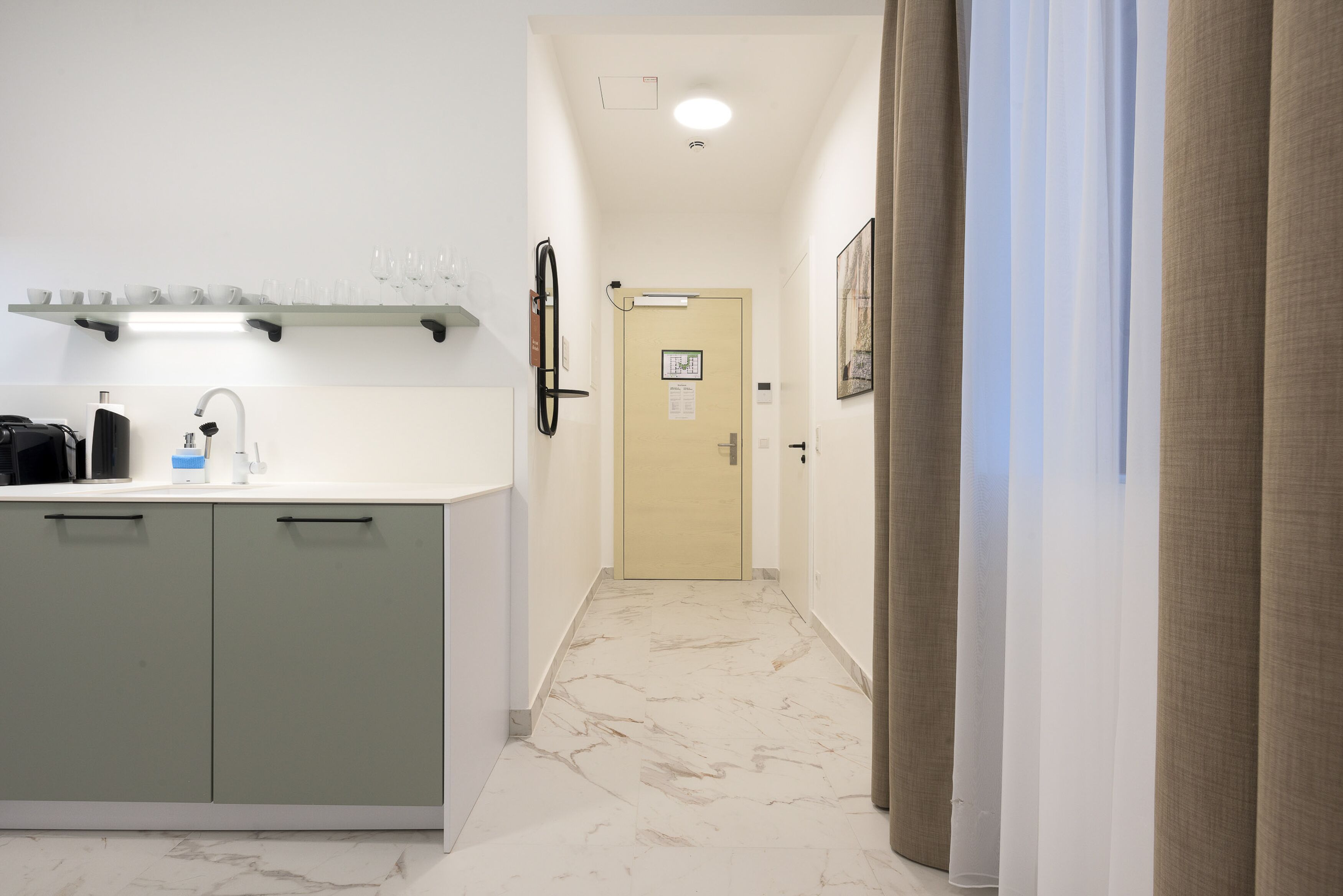 A narrow hallway with a kitchen area on the left, featuring cabinets and a countertop with a sink. A door at the end of the hallway leads to another room.