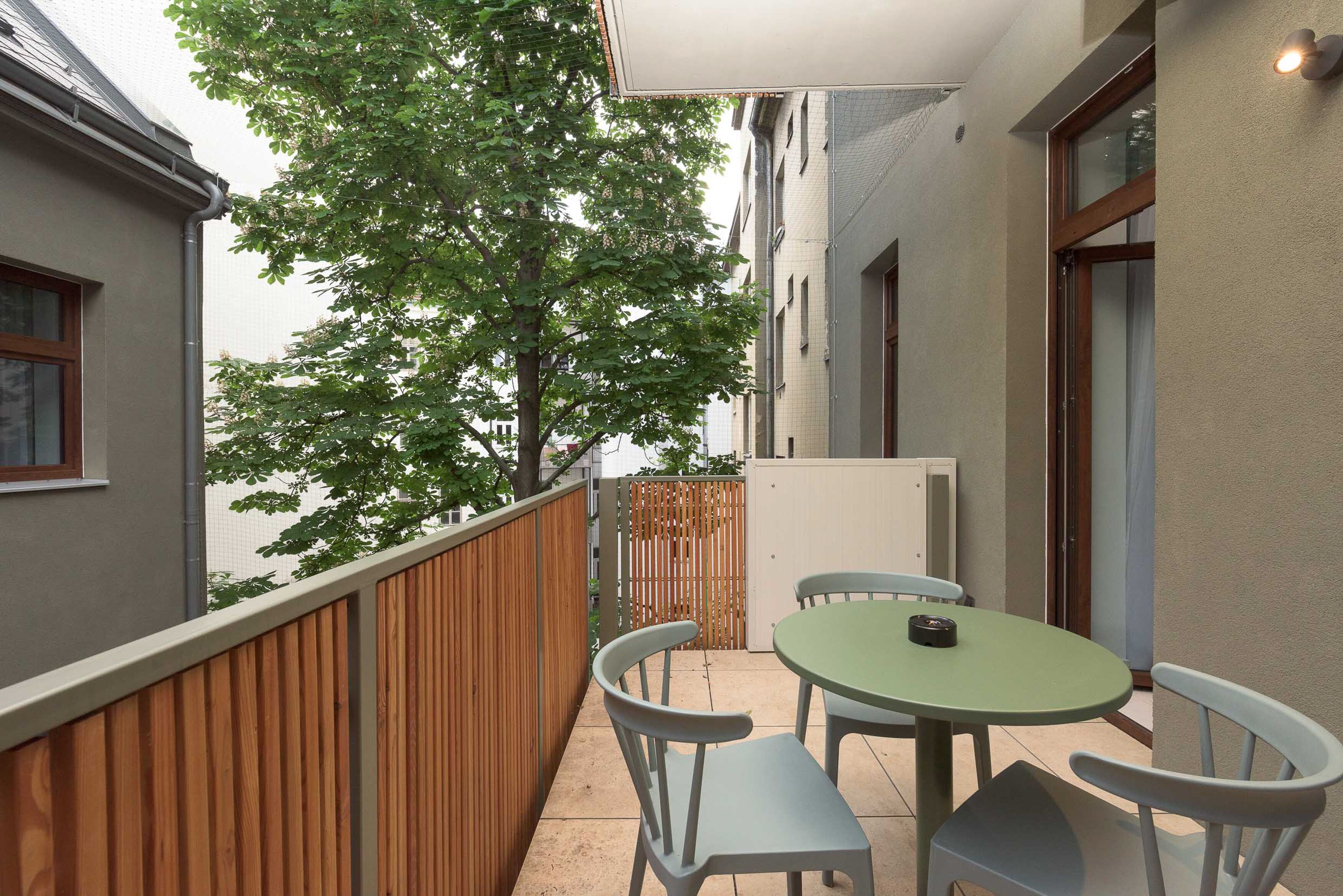A balcony with a wooden fence, a round table with two chairs, and a view of a tree and buildings in the background.