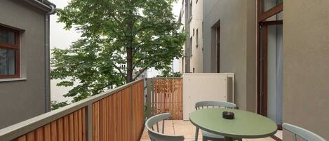 A balcony with a wooden fence, a round table with two chairs, and a view of a tree and buildings in the background.